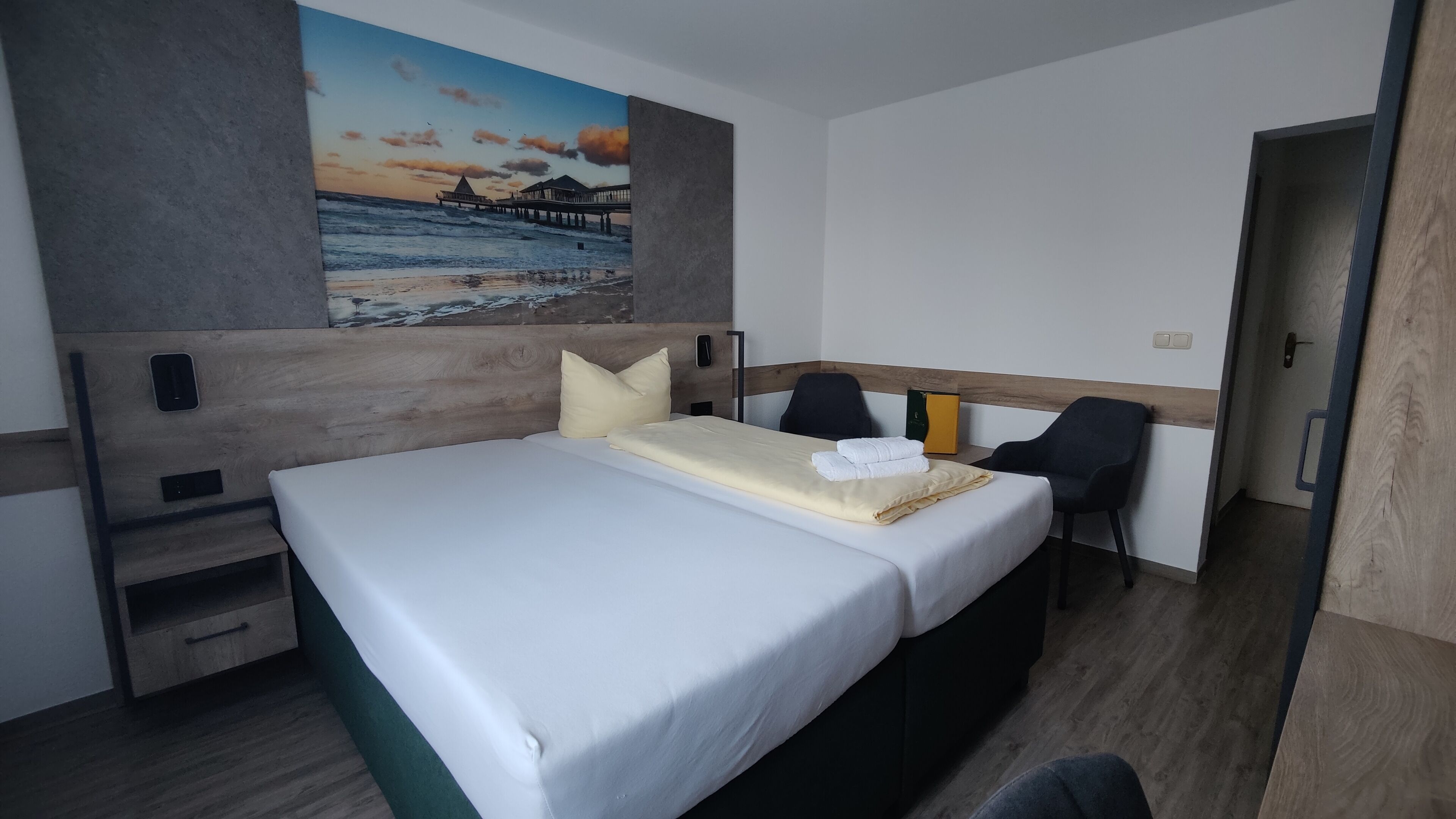 A hotel room with a bed, a desk, a chair, and a painting of a beach scene.