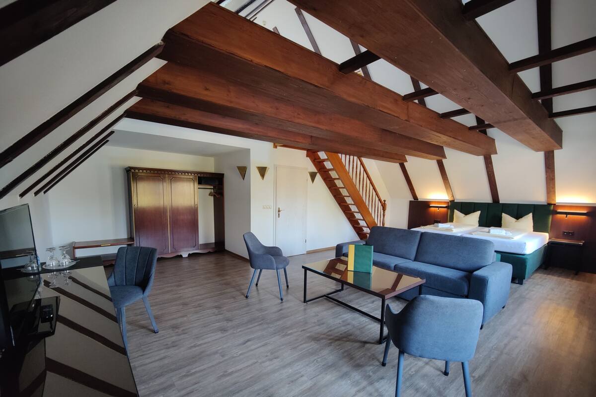 Un espacioso dormitorio tipo loft con techo alto, vigas de madera a la vista, una cama grande, zona de estar con sofá y sillones, y una escalera que sube a un nivel superior.