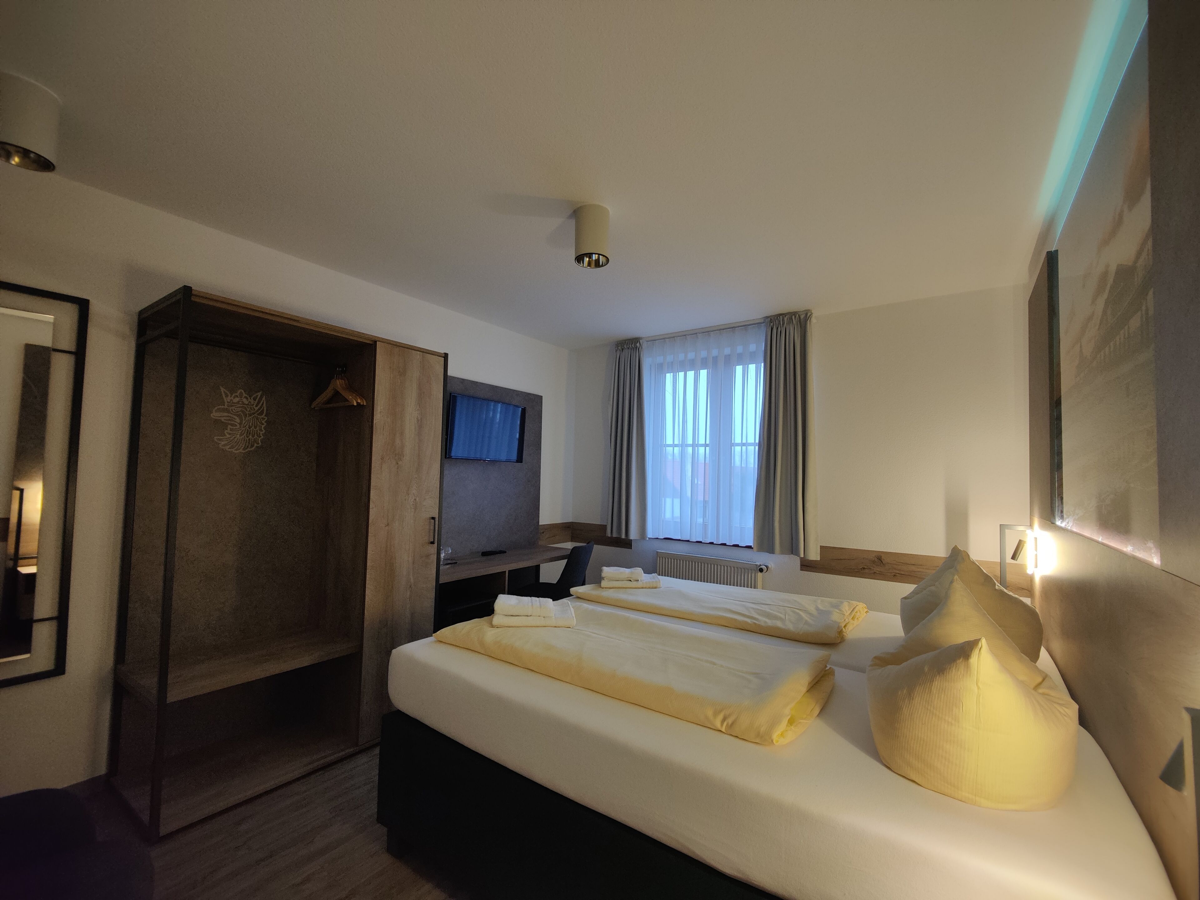 Una habitación de hotel con cama, escritorio, armario, televisor y ventana con cortinas.
