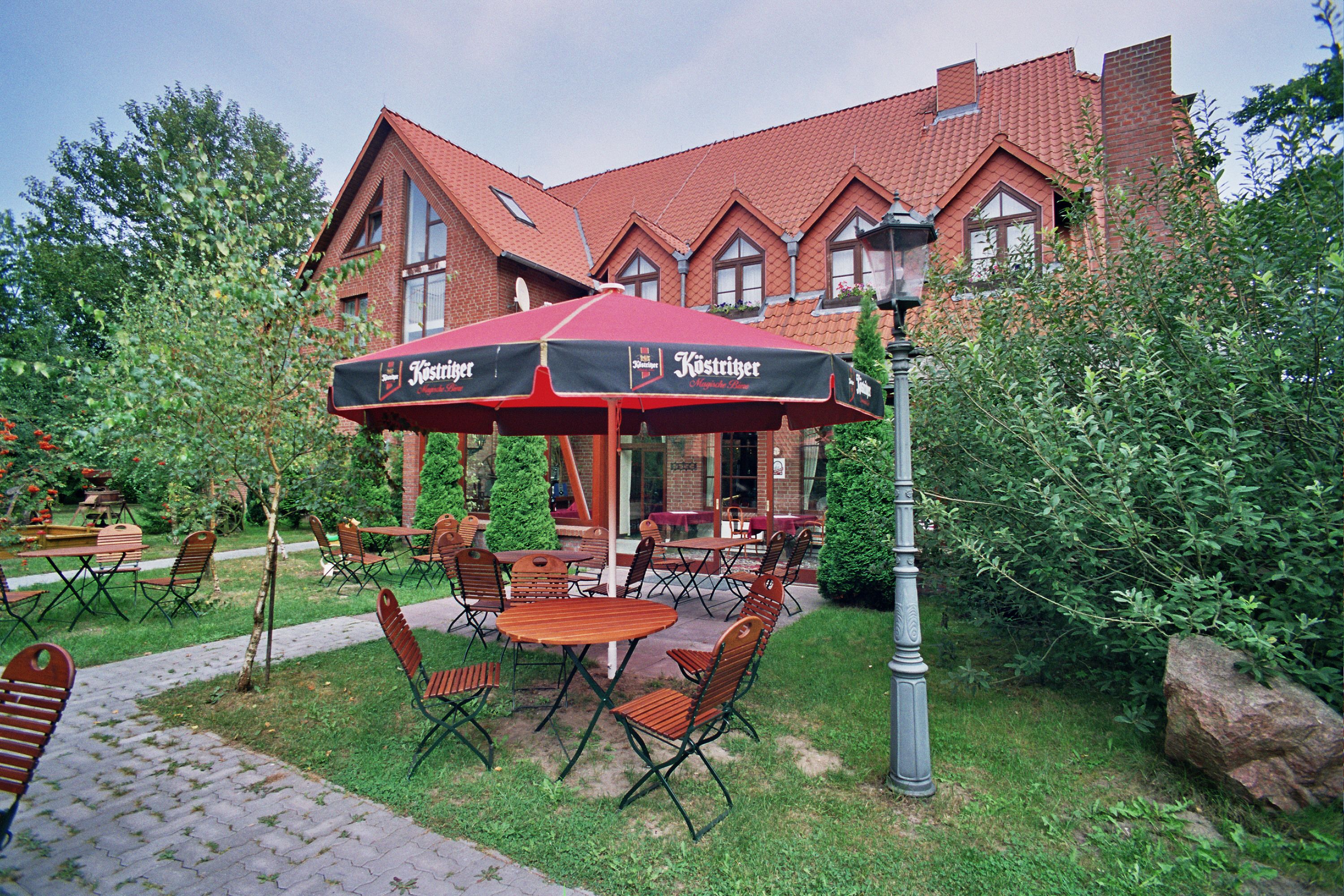 An outdoor seating area with tables and chairs, a red umbrella, and a brick building in the background.