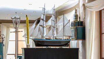 Un modelo de barco exhibido en una habitación con techo alto, ventanas amplias y decoración clásica, incluyendo una lámpara y cortinas.