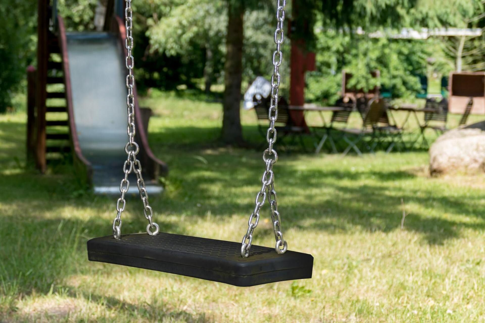 A black swing suspended by metal chains in a grassy park area with playground equipment in the background.