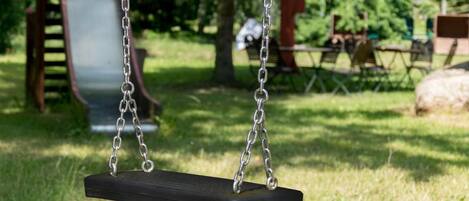 A black swing suspended by metal chains in a grassy park area with playground equipment in the background.