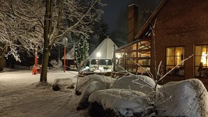 Un espacio exterior cubierto de nieve con un edificio de ladrillo, una estructura de madera con techo inclinado y un árbol nevado.