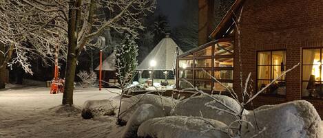 Un espacio exterior cubierto de nieve con un edificio de ladrillo, una estructura de madera con techo inclinado y un árbol nevado.
