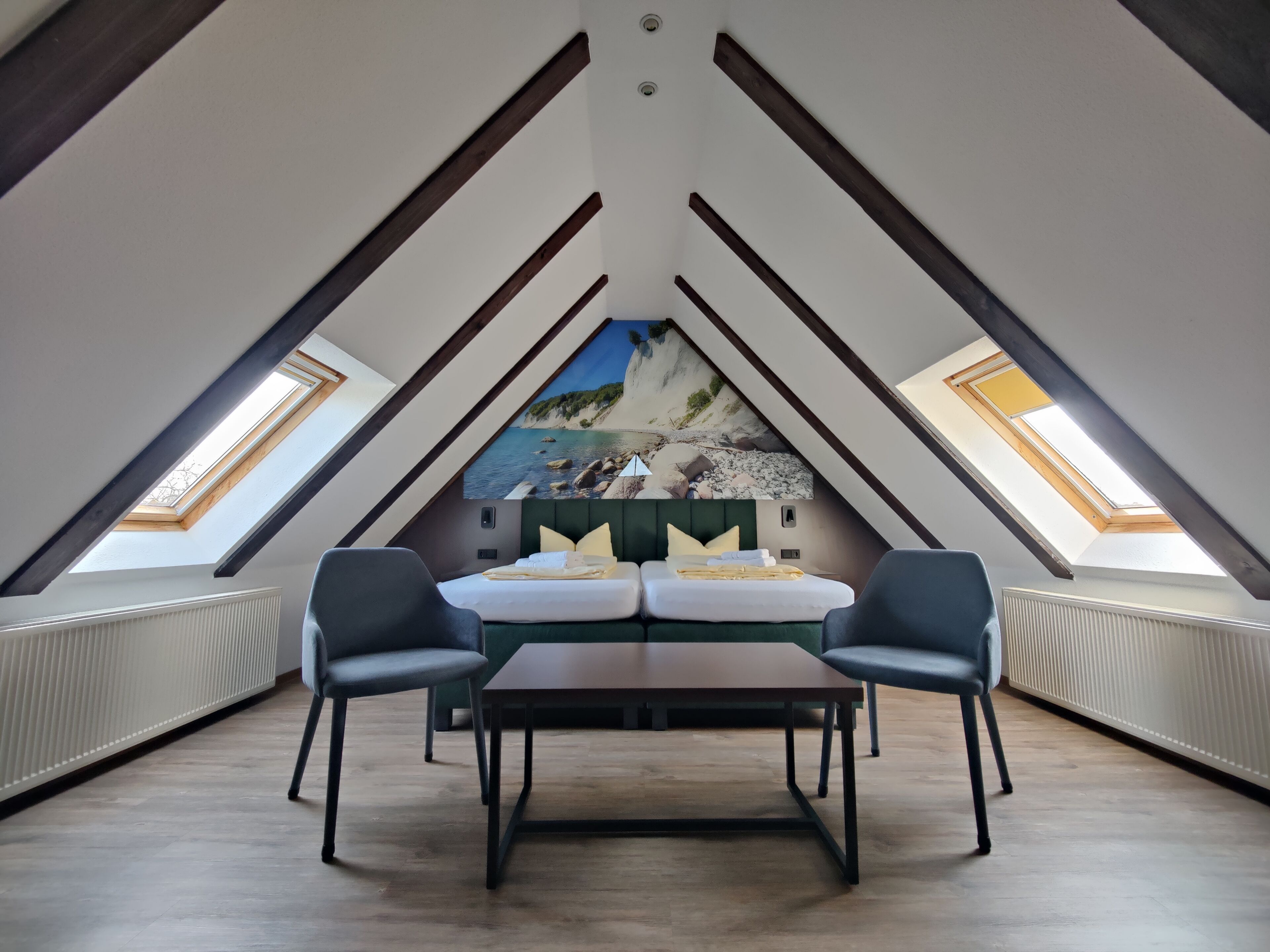 Een kamer met een schuine plafond, een bed met kussens, twee stoelen, een tafel en een muurschildering van een strand.