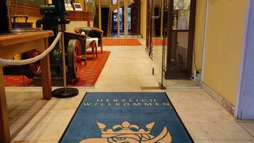 A hotel lobby with a blue carpet featuring a bird design and the text "Herzlich Willkommen" (Welcome). There are signs indicating Michelin and Holiday Inn Express awards, a reception area with a counter and a coffee machine, and a glass door leading to a room.