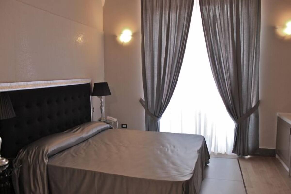 A bedroom with a large bed, a headboard, a bedside lamp, and curtains.