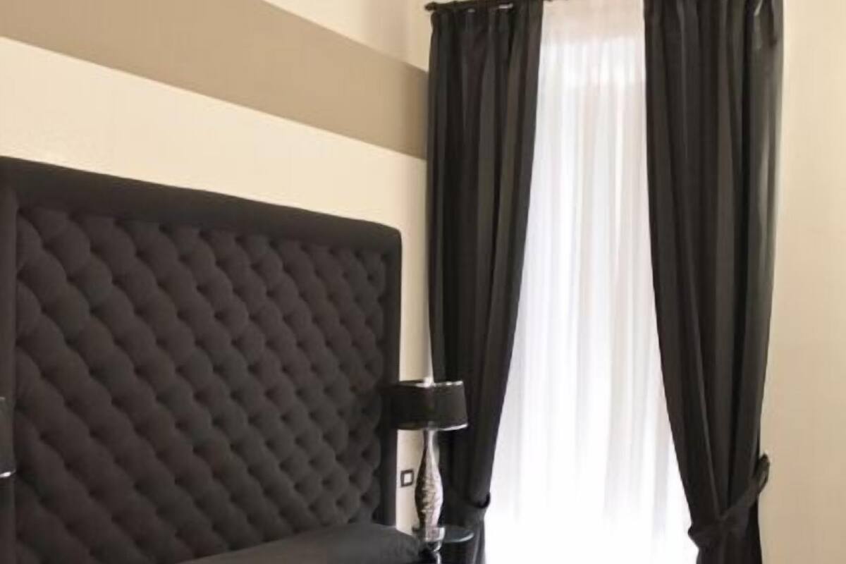 A bedroom with a large bed, a tufted headboard, a bedside table with a lamp, and dark curtains.