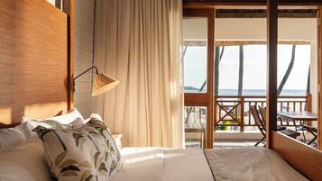 A bedroom with a large bed, a wooden headboard, a hanging lamp, and a view of the beach through an open door.