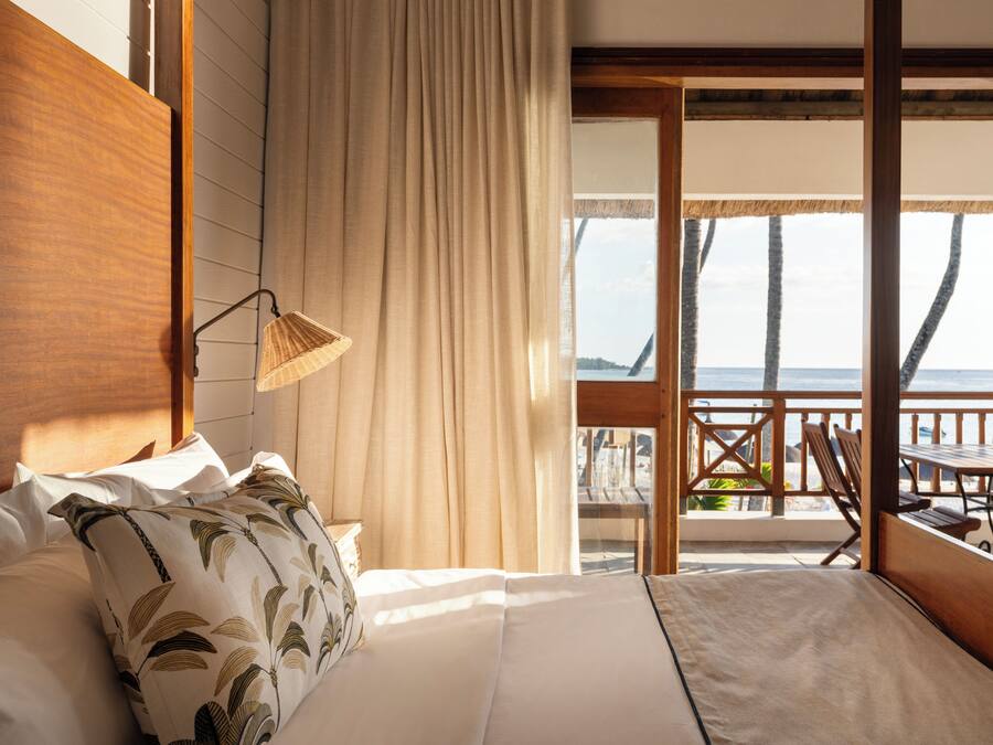 A bedroom with a large bed, a wooden headboard, a hanging lamp, and a view of the beach through an open door.