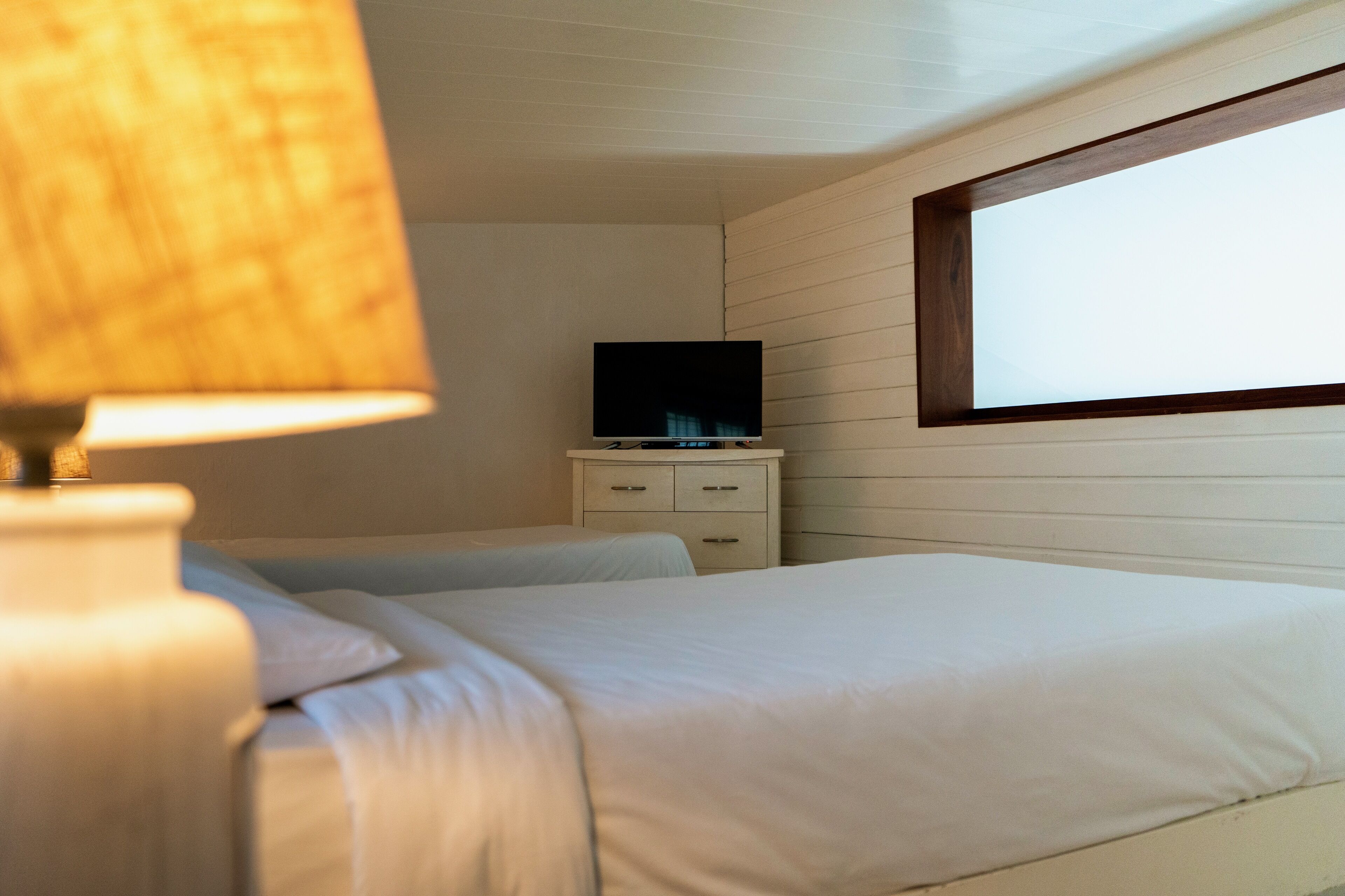 Un dormitorio con dos camas, una mesita de noche con televisor y una ventana que proporciona luz natural.