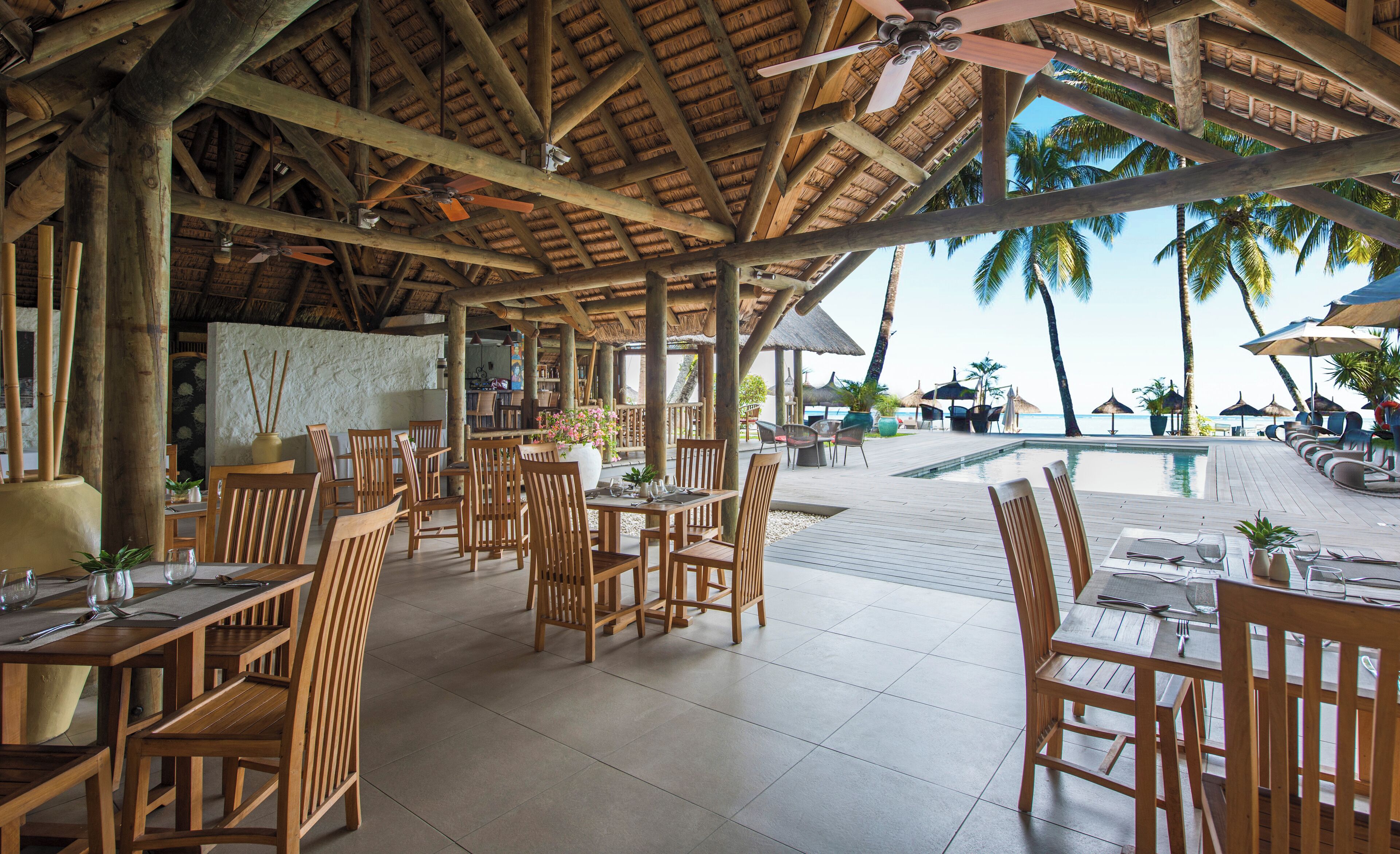 A wooden deck with tables and chairs, overlooking a pool and beach.