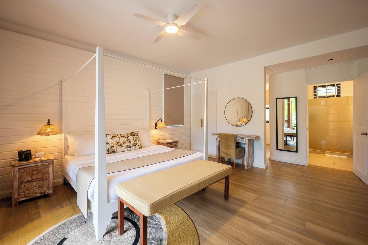 A bedroom with a four-poster bed, a bench, a bedside table with a lamp, a mirror, and a view into the bathroom.