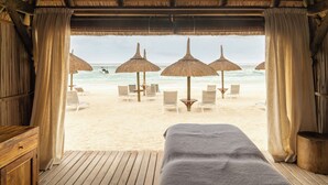 A beach view from a room with a bed, wooden floor, and curtains. Outside, there are beach chairs, umbrellas, and the ocean. - Constance Sakoa Boutik (Trou aux Biches)