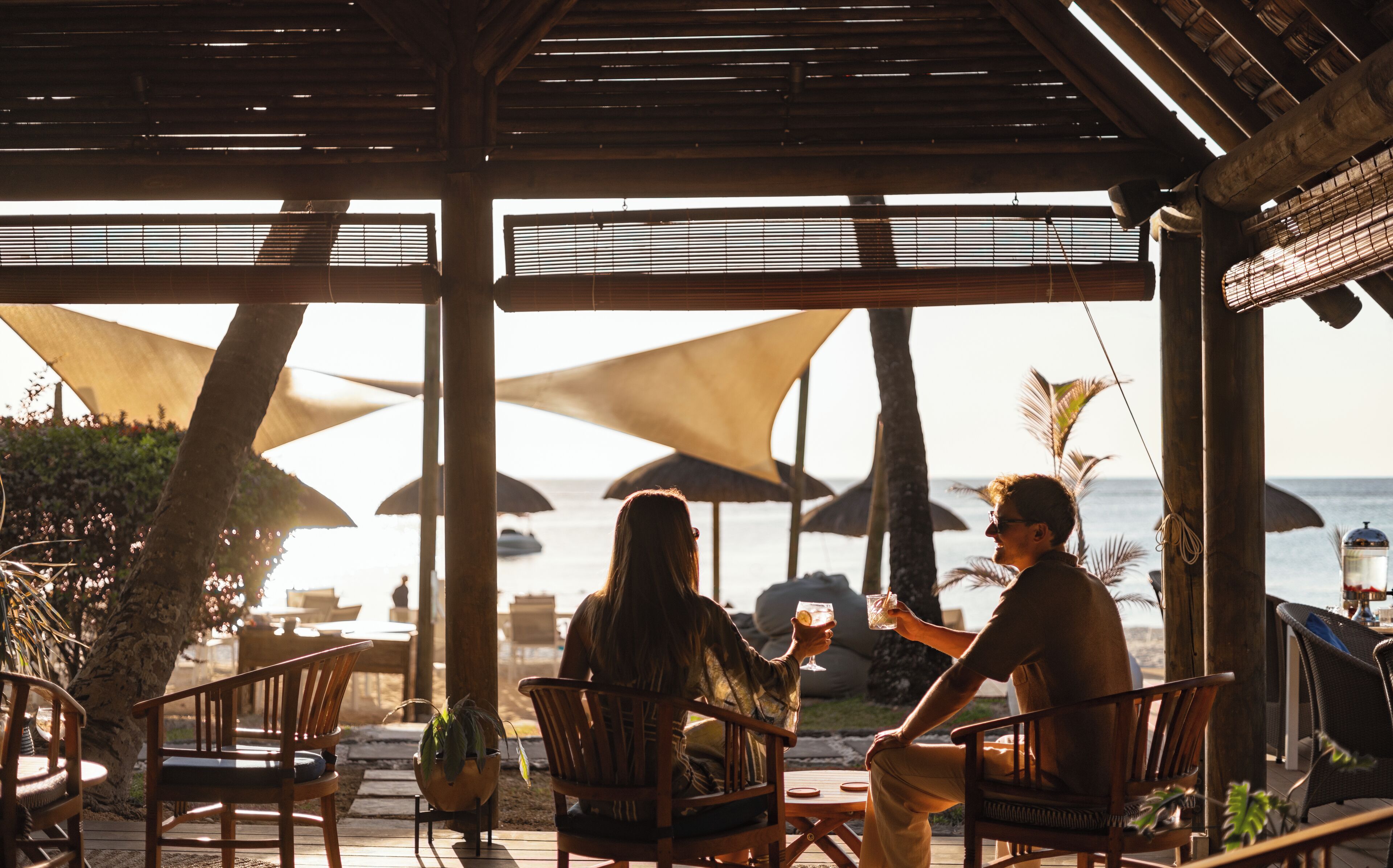 A covered outdoor seating area with wooden furniture and umbrellas, overlooking a beach.