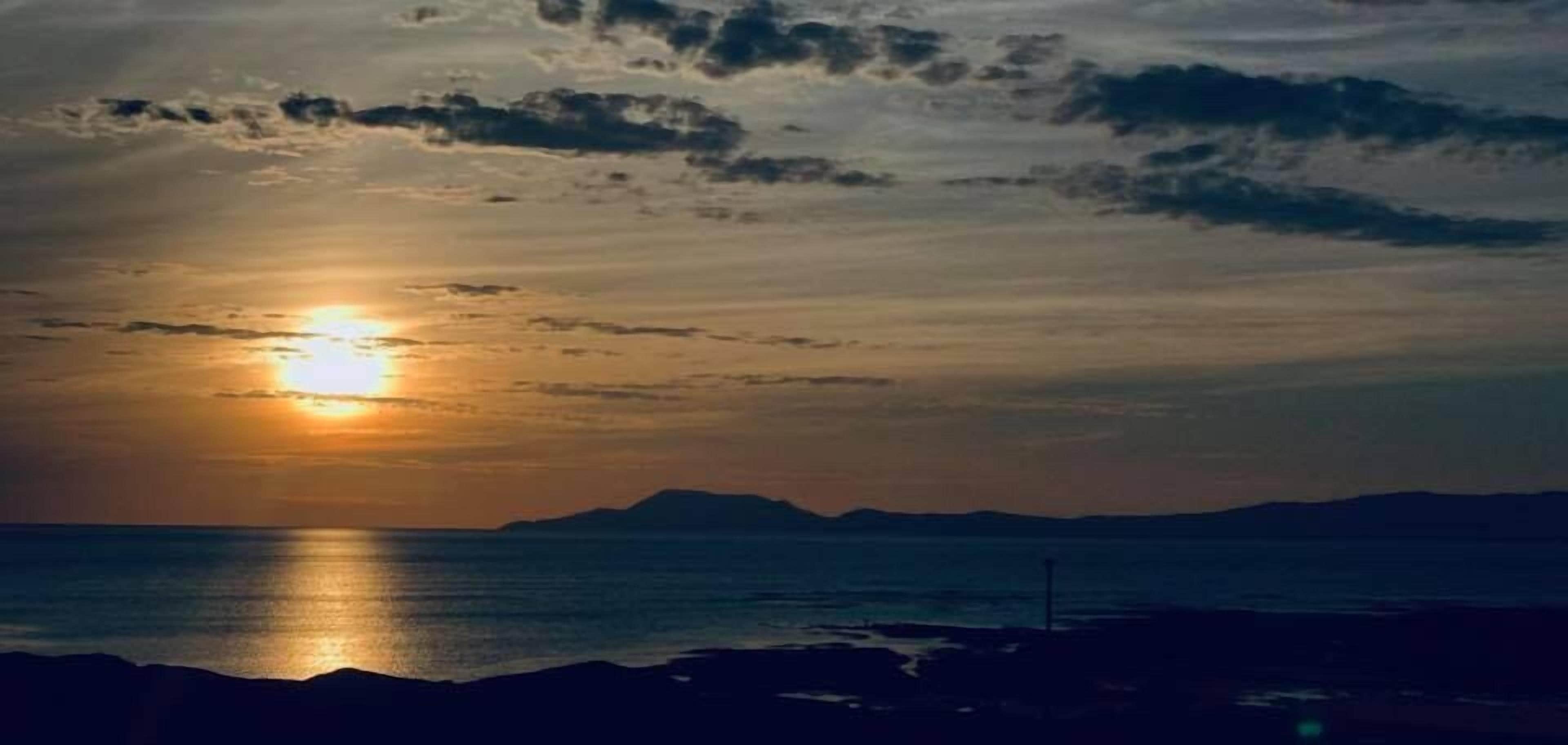 Un coucher de soleil sur un paysage côtier, avec les silhouettes de montagnes et une mer calme.