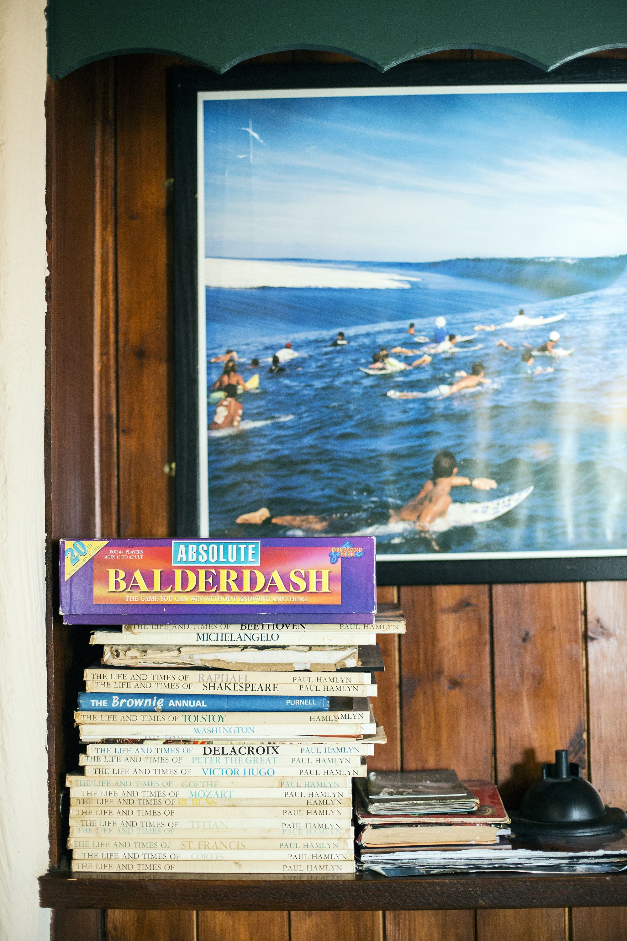 A stack of books with a framed picture of surfers on the wall behind them.