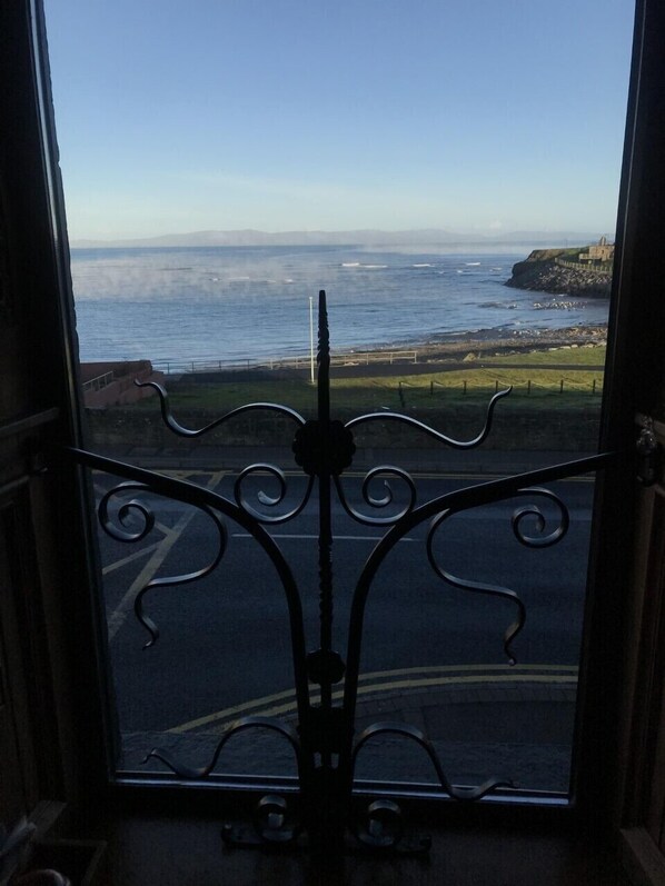 Vista a través de una ventana con una reja de hierro decorativa, con vista a una zona costera con cielo despejado y mar tranquilo.
