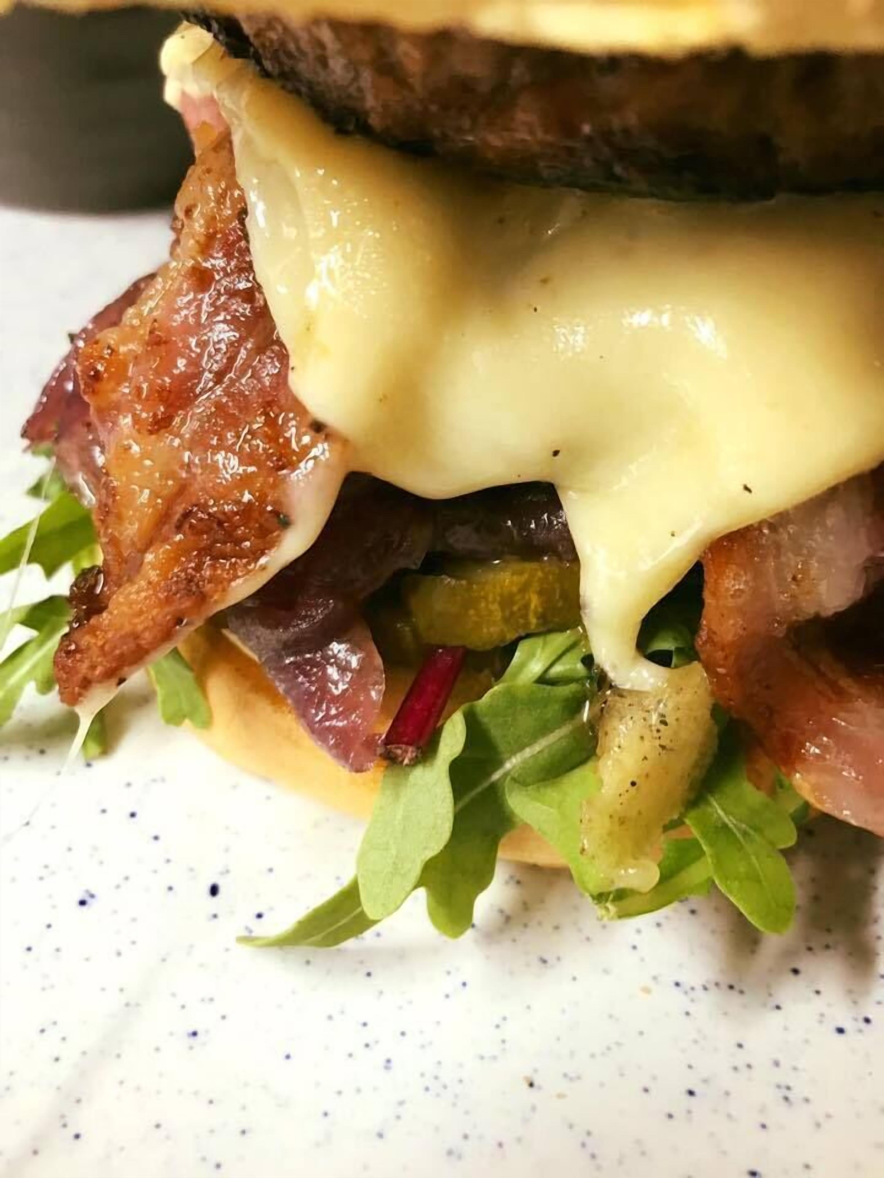 Primer plano de un sándwich con queso derretido, tocino y pepinillos sobre un plato blanco.