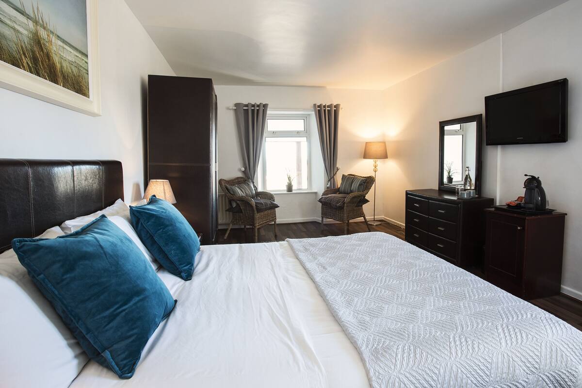 Habitación de hotel con una cama grande, dos sillas, un aparador, televisor y ventana con cortinas.