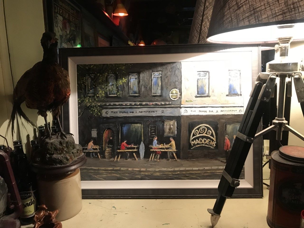A framed artwork of a street scene with tables and chairs outside a building, a tripod with a camera, and a potted plant.
