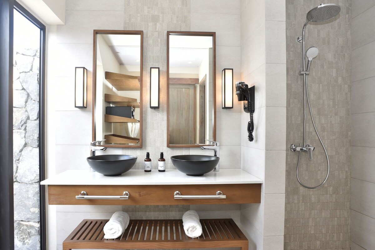Een moderne badkamer met twee grote spiegels, een zwarte wastafel, een houten wastafelmeubel, een handdoekhouder en een douche met een handdouche.