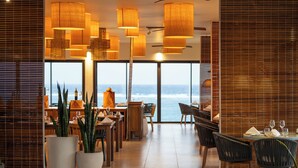 Un restaurante con amplios ventanales con vista al océano, muebles de madera y lámparas colgantes.