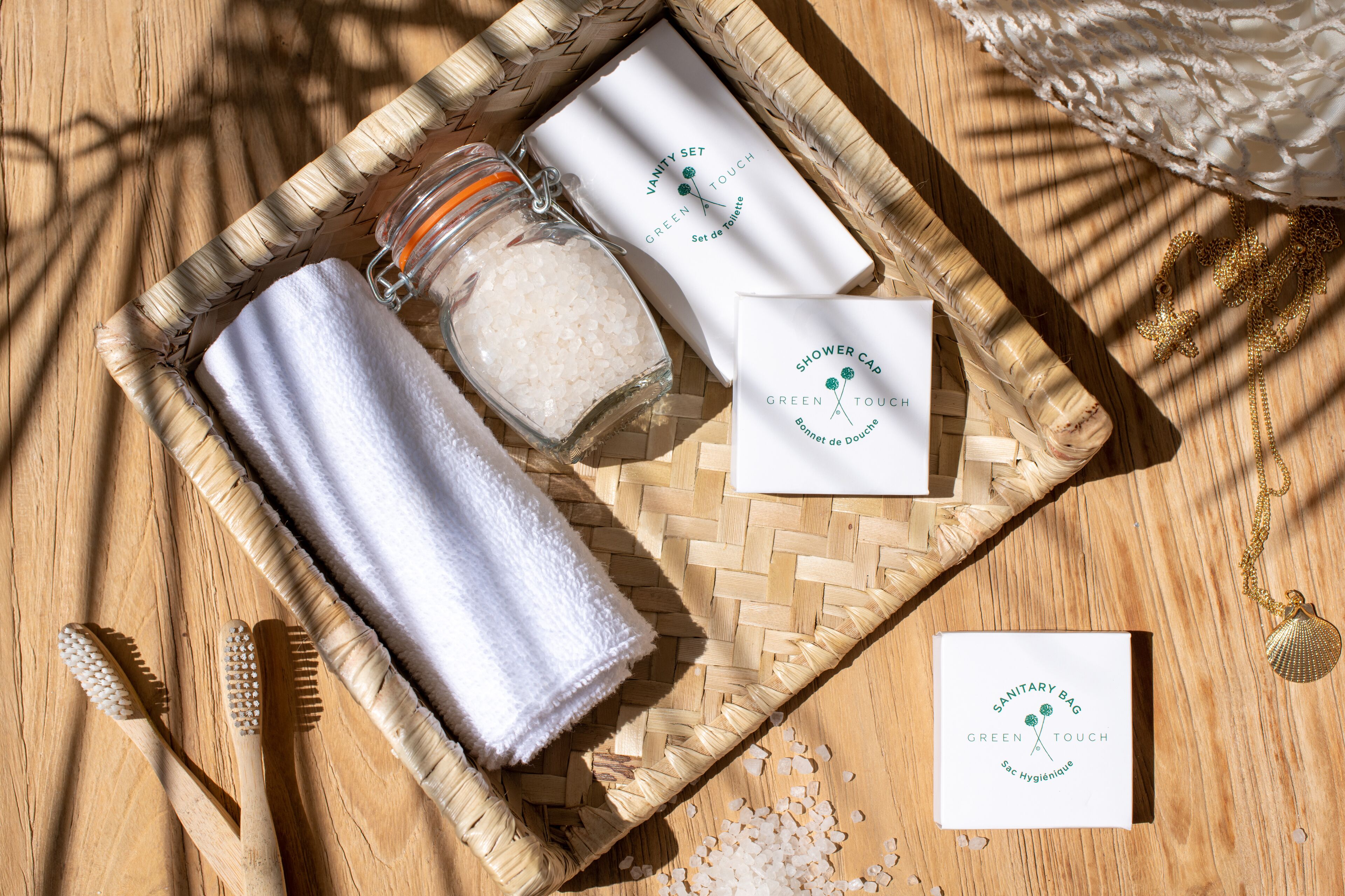 A wooden tray with a white towel, a glass jar of bath salts, a toothbrush, and a card labeled "SANITARY BAG GREEN TOUCH."