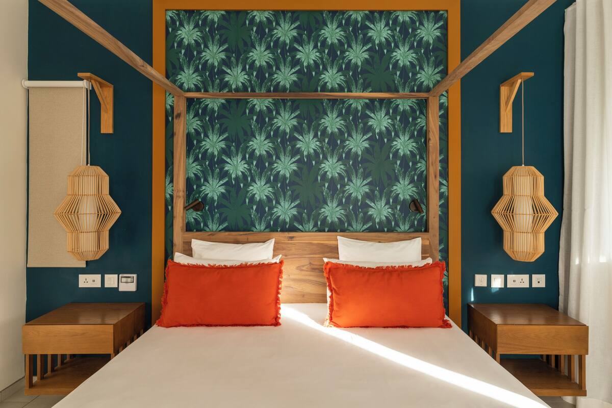 Une chambre à coucher avec une tête de lit en bois, deux coussins rouges et un mur orné d’un motif de palmiers.