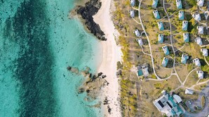 Vista aérea de un resort costero con varias construcciones tipo cabaña, una playa de arena y aguas turquesas cristalinas.