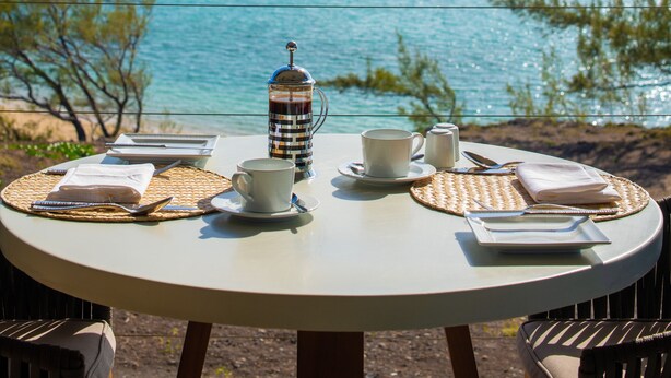 Una mesa redonda con vistas al océano, una cafetera, tazas y un cuaderno.