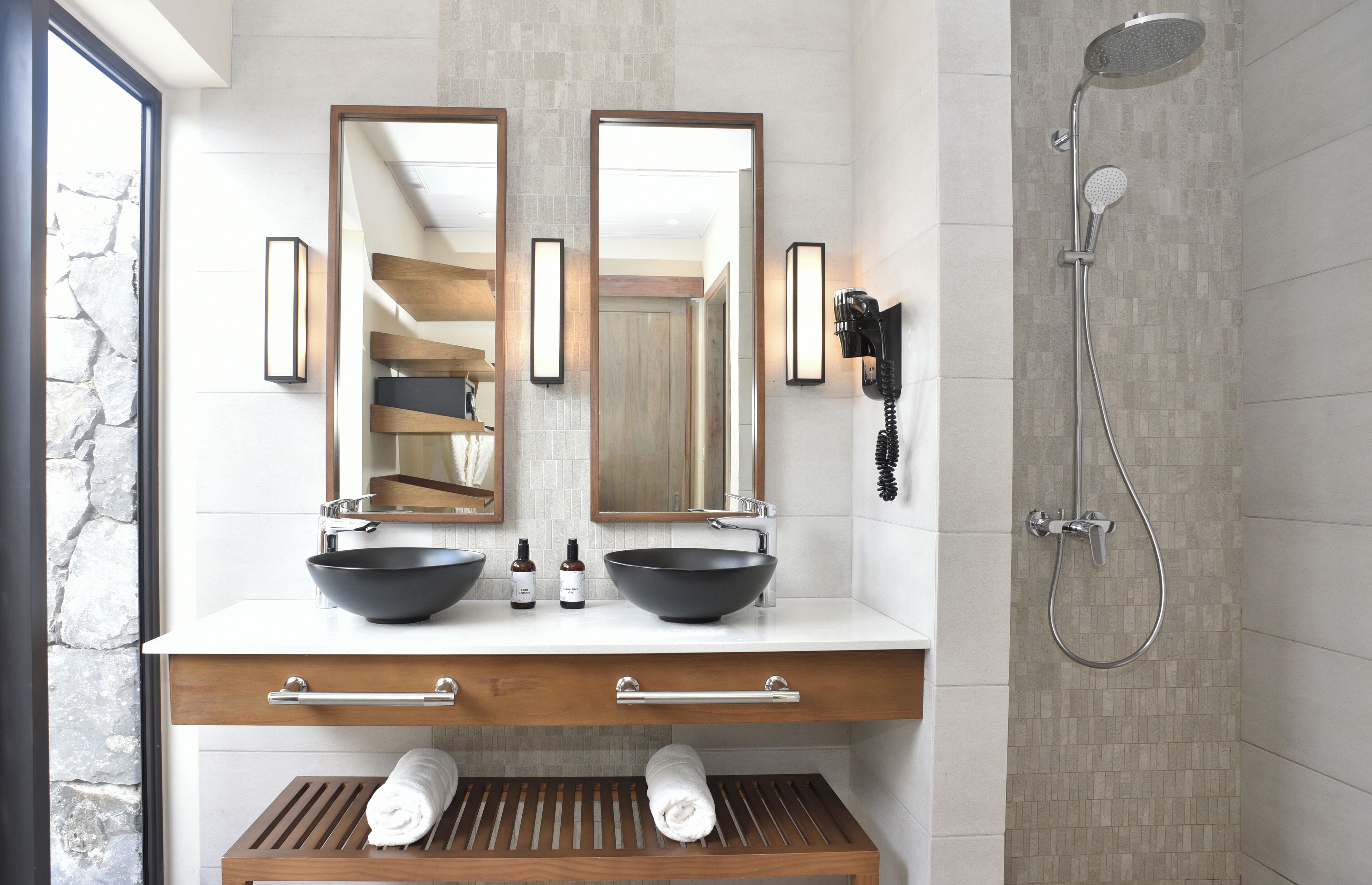 A modern bathroom with two large mirrors, a black vessel sink, a handheld showerhead, and a towel rack.