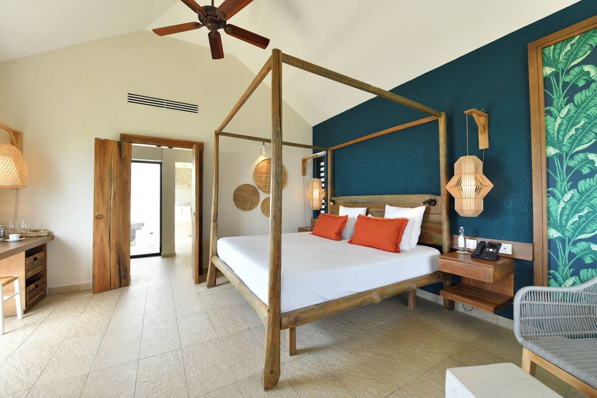 Un lit à baldaquin avec du linge de lit blanc et des coussins orange, une table de chevet en bois, une chaise en osier et un ventilateur de plafond.