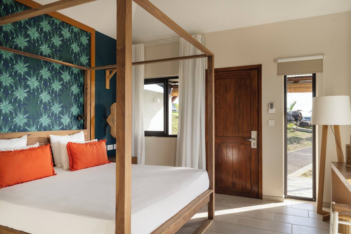 Un lit à baldaquin avec des oreillers blancs et orange, une tête de lit en bois, un mur bleu foncé orné d’un motif de palmier, une porte en bois et une lampe sur une table d’appoint.