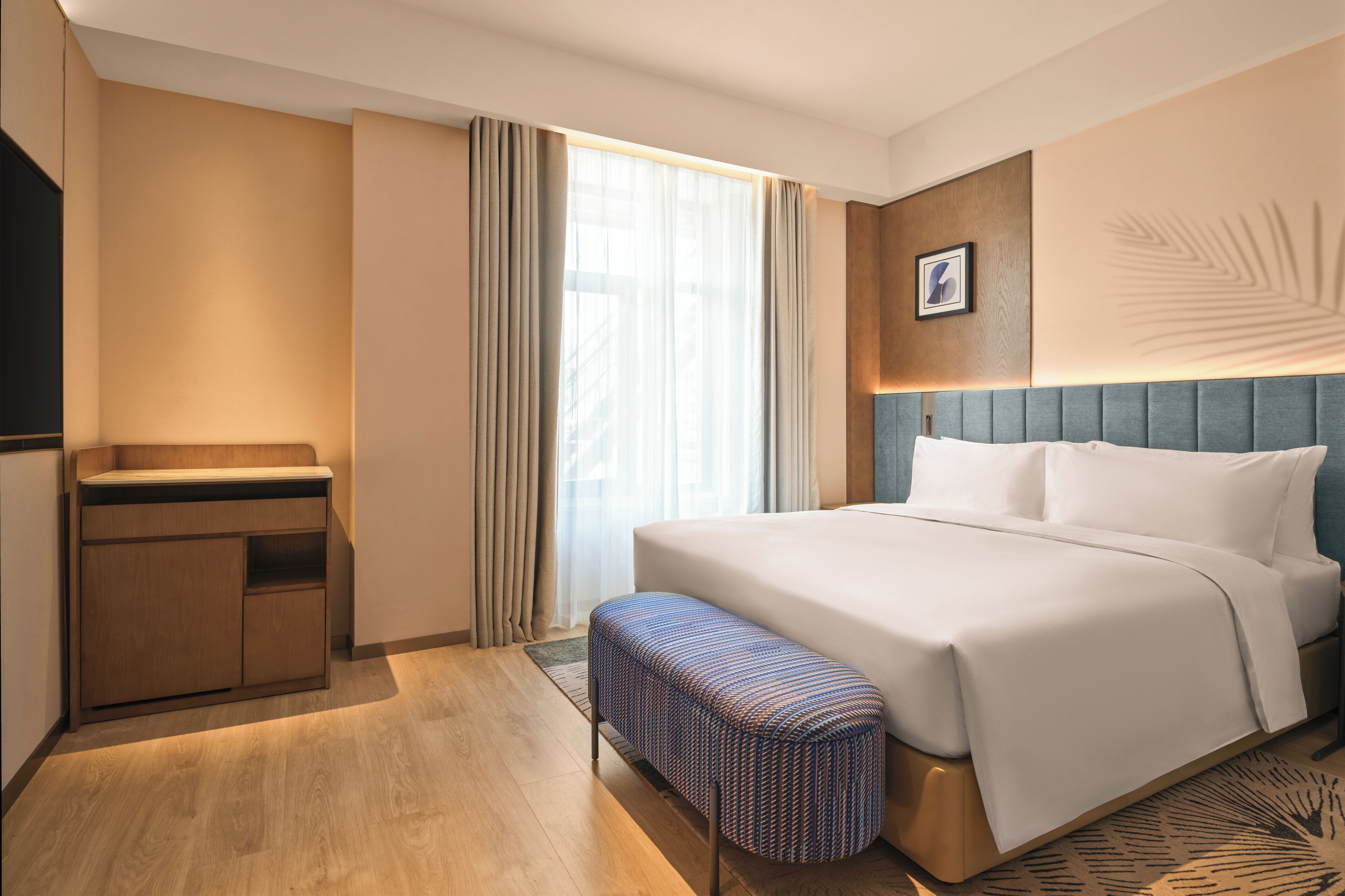 Habitación de hotel con cama, una banca, escritorio y un amplio ventanal con cortinas.