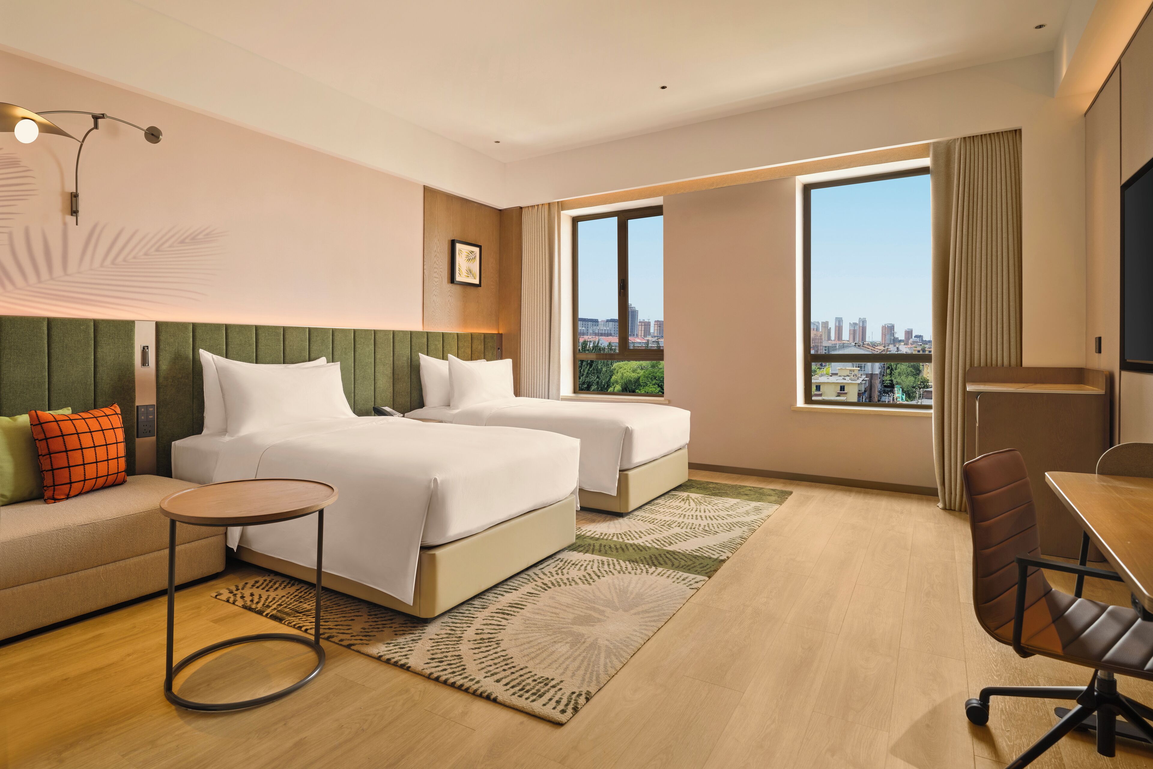 A hotel room with two beds, a sofa, a desk, and a large window with a city view.