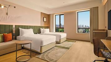 A hotel room with two beds, a sofa, a desk, and a large window with a city view.
