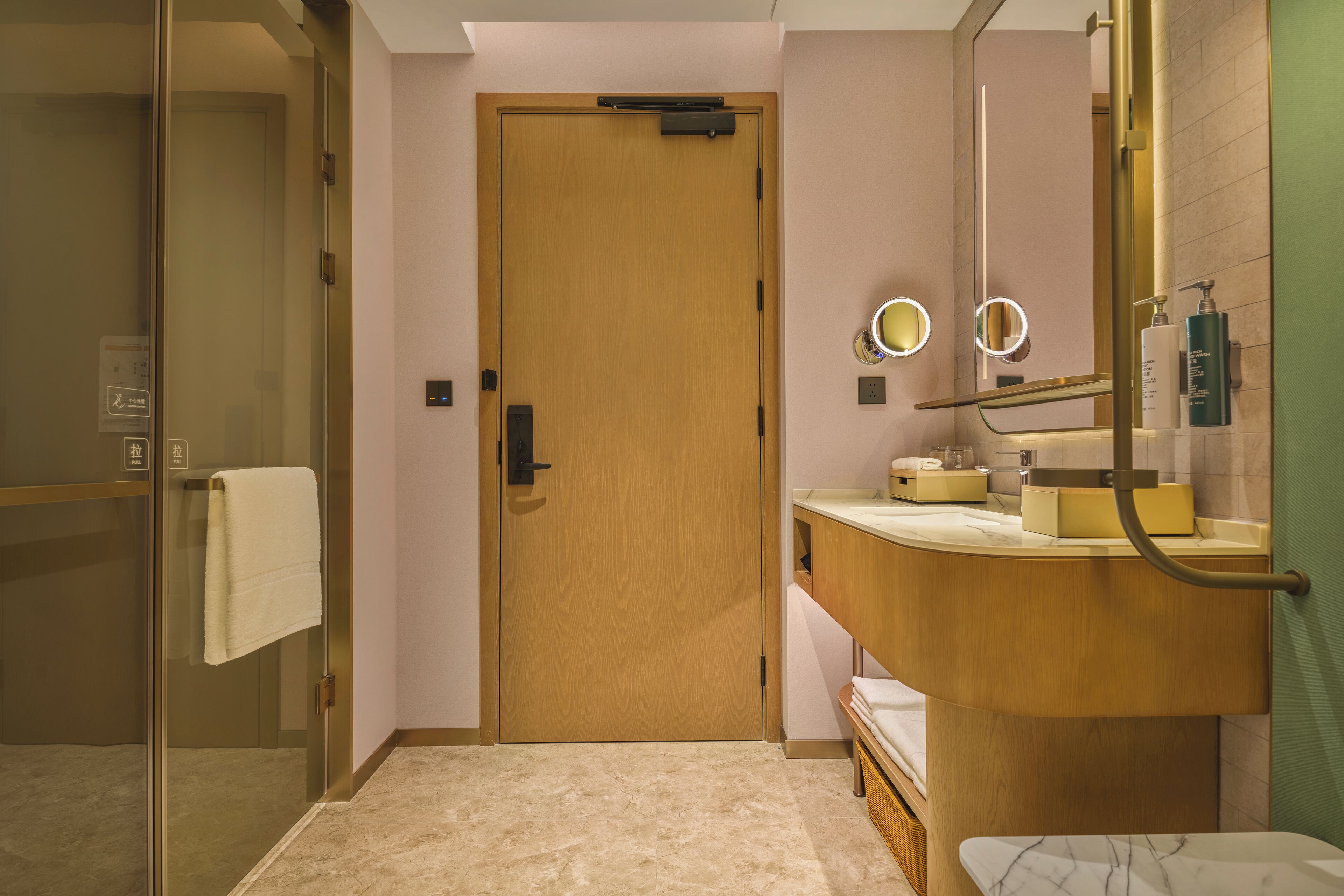 Un baño moderno con puerta de madera, cabina de ducha de vidrio, lavamanos de mármol y espejo con luz circular.