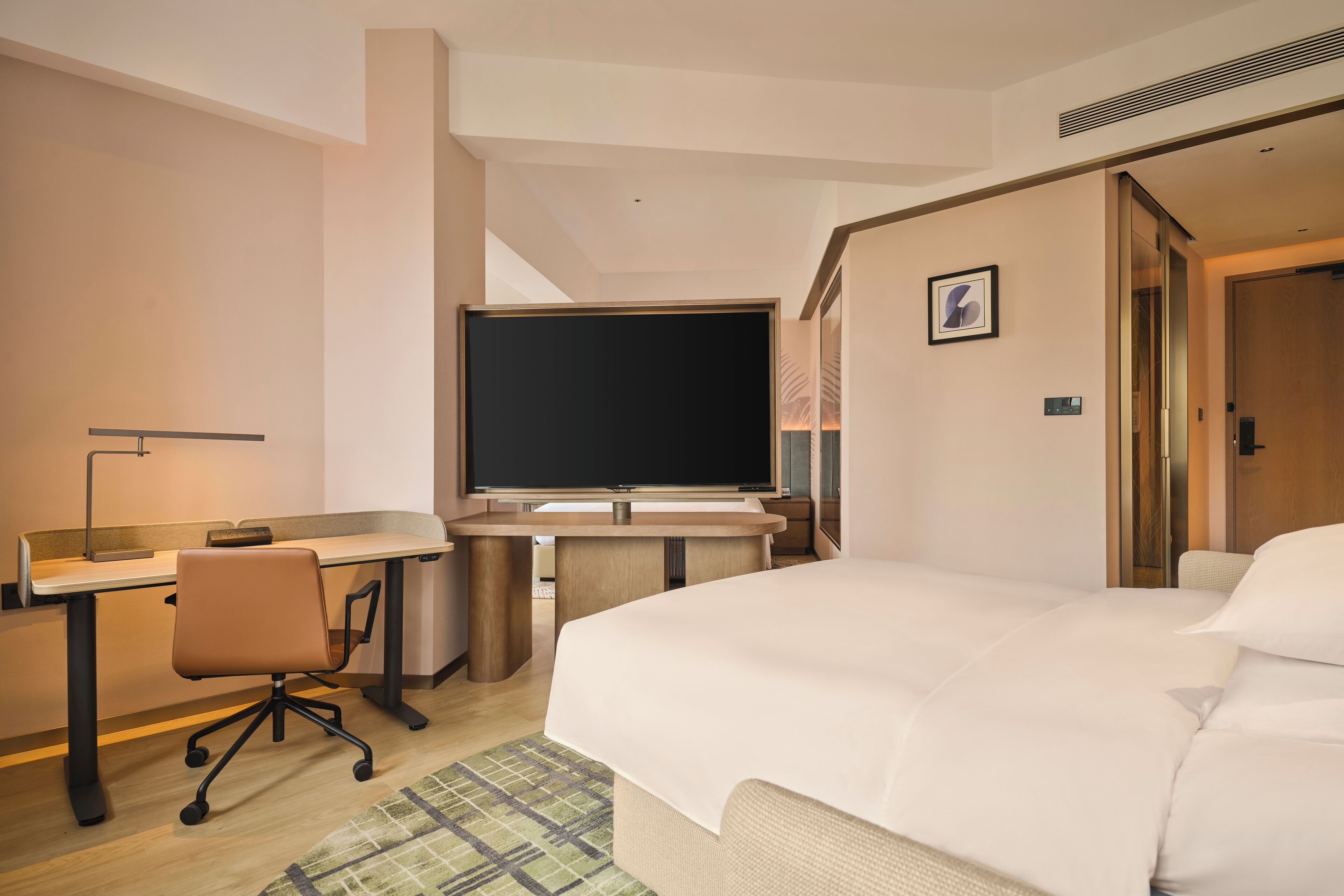 A hotel room with a bed, a desk with a chair, a flat-screen TV, and a door.