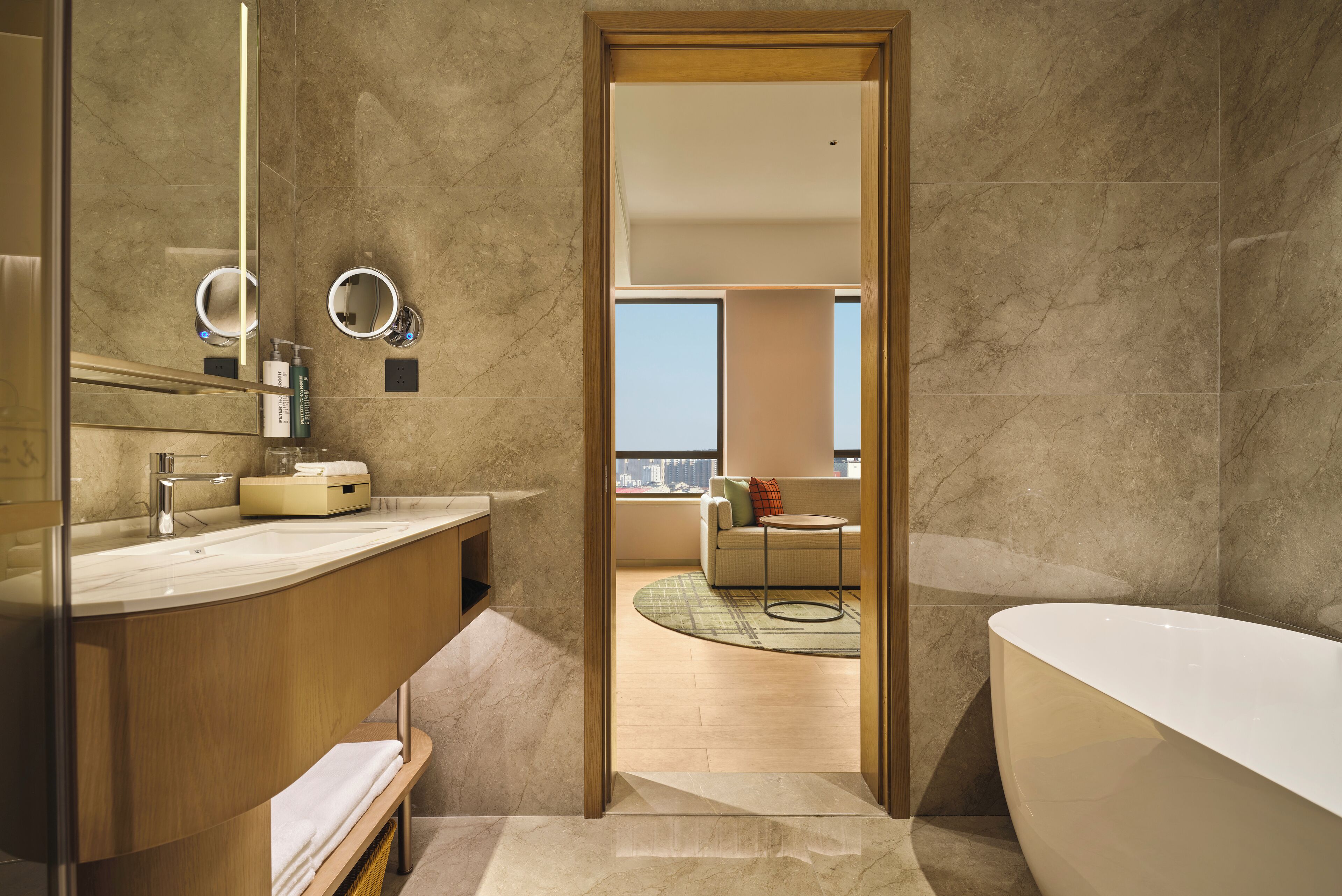 A modern bathroom with a large bathtub, a vanity with a mirror, and a view of a living room with a sofa and a cityscape.