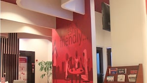 A modern lobby with a red wall featuring a 'fun, fresh & friendly' sign, a reception desk, and a magazine rack.