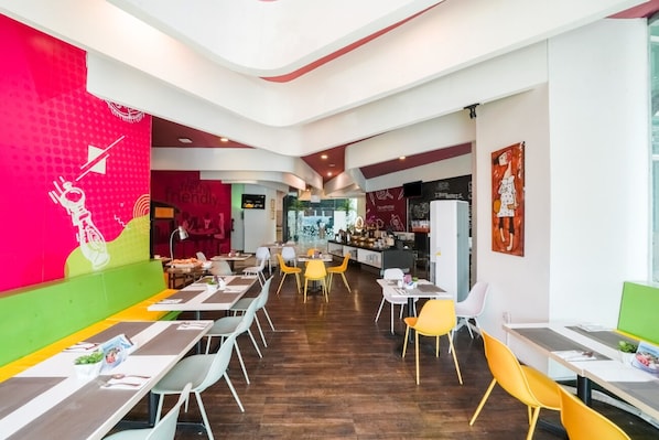 Un restaurant au décor moderne, avec un long comptoir bordé de chaises jaunes, un mur rouge et vert orné d’un personnage de dessin animé, et un bar situé au fond de la salle.