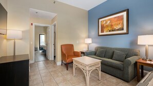 Ein Hotelzimmer mit einer beigen Wand, einer blauen Akzentwand, einem gerahmten Gemälde, einem grün gestreiften Sofa, einem orangefarbenen Sessel, einem weißen Tisch und zwei Lampen.