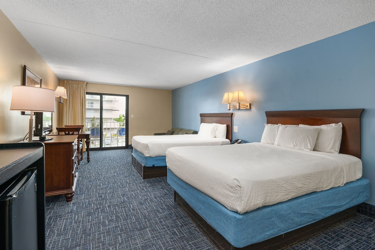 A hotel room with two beds, a desk, a chair, a lamp, and a view of the outside.