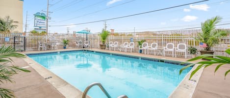 A swimming pool with a metal ladder, surrounded by white chairs and potted plants, with a sign for Coastal Palms Beach Hotel in the background.