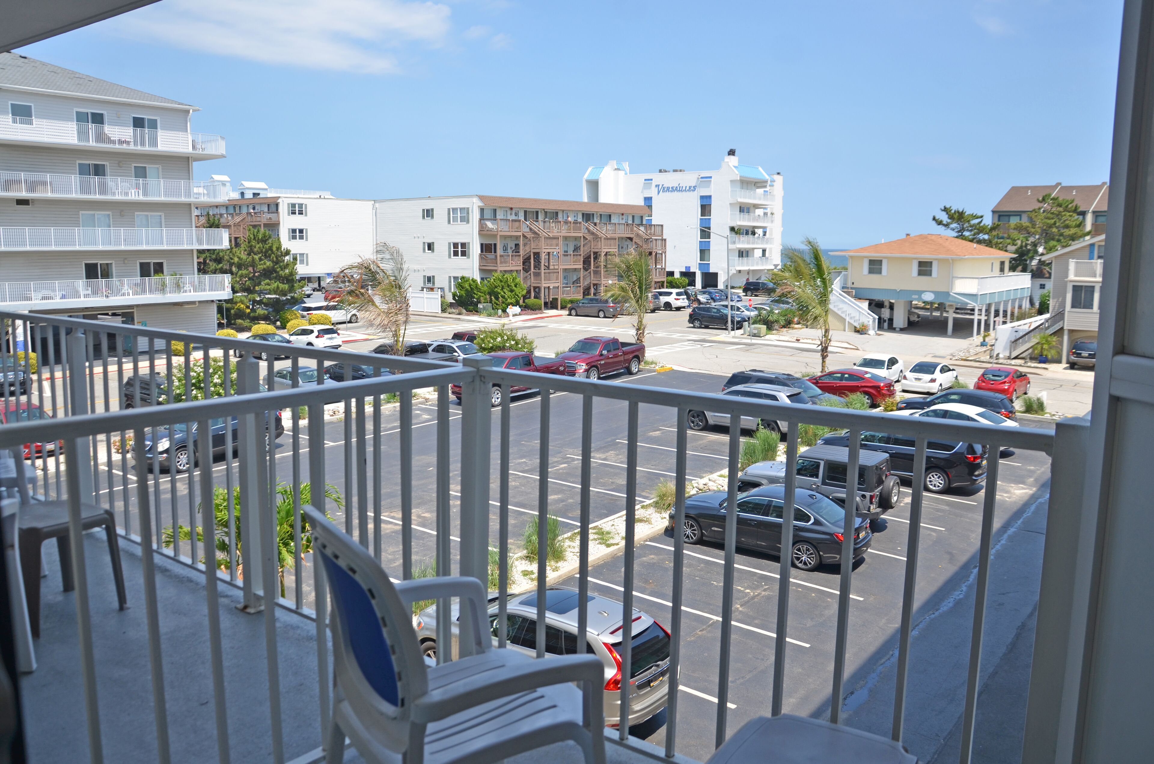 A view from a balcony overlooking a parking lot with several cars, a building with a blue and white facade, and a clear sky.
