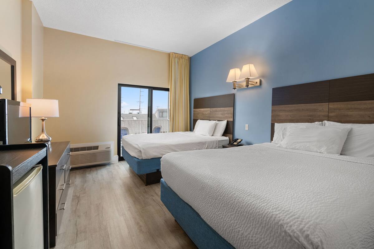 A hotel room with two beds, a desk, a lamp, and a view of the outside.