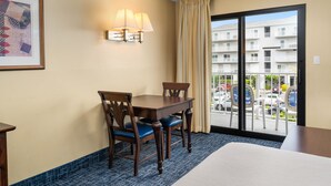 Ein Hotelzimmer mit Balkon, einem Esstisch mit Stühlen, einem gerahmten Bild an der Wand und einer Lampe.