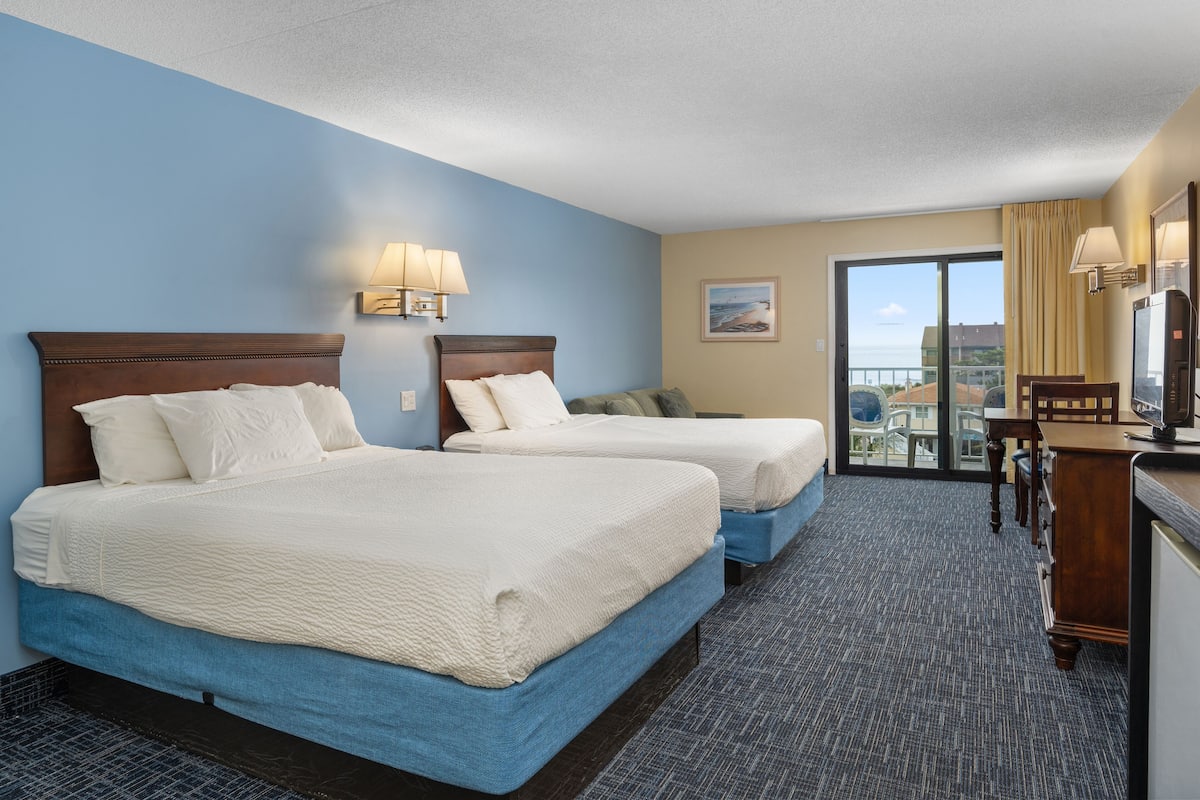 A hotel room with two beds, a desk, a chair, and a balcony with a view.